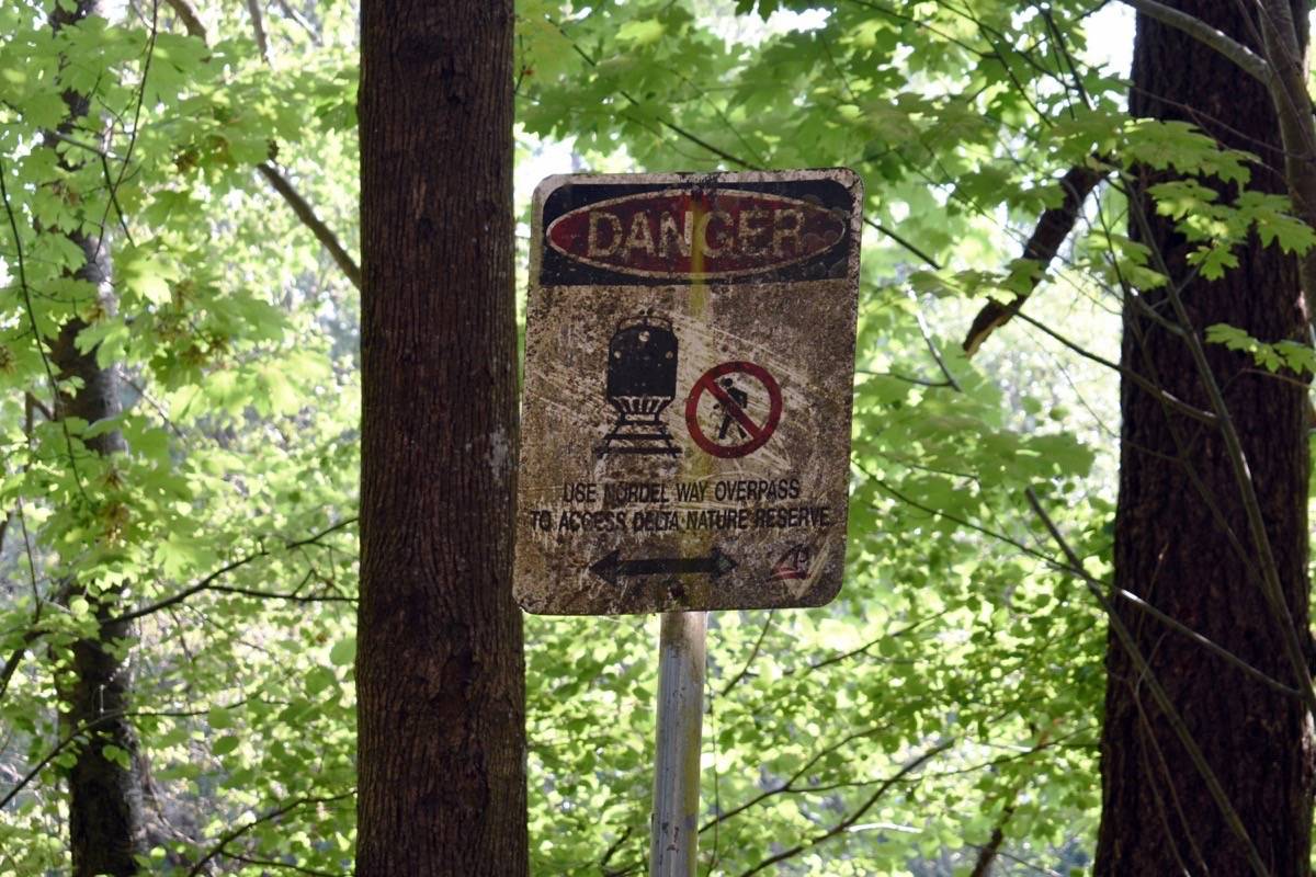 A weathered sign at the foot of the trail leading from Monroe Drive to the North Delta Greenway advises people use the Nordel Way overpass to access the Delta Nature Reserve. The trail and stairs to the unsanctioned rail crossing are a few metres to the left. (James Smith photo)