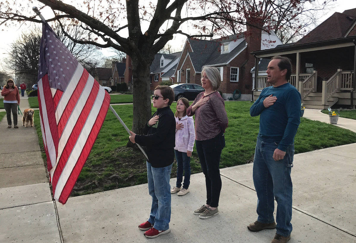 In this photo taken April 7, 2020, Zach Stamper holds the U.S. flag while his sister Juliette and parents Jennifer and Tim recite the Pledge of Allegiance in the driveway of their home, as next door neighbor, Ann Painter, left, participates in Kettering, Ohio. The Pledge has become a morning ritual in their neighborhood since schools closed due the COVID-19 threat. (AP Photo/Mitch Stacy)