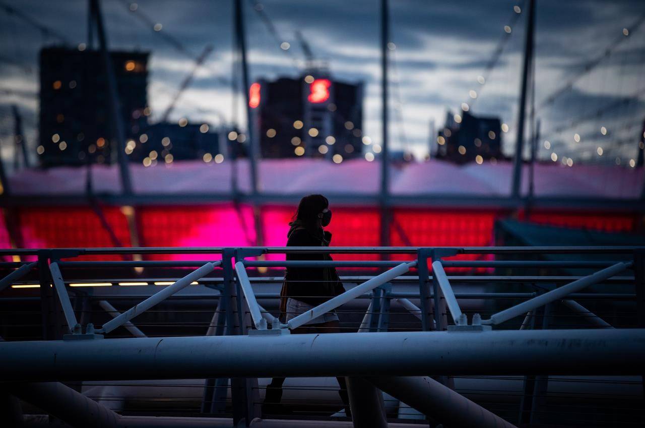 A person wearing a face mask to curb the spread of COVID-19 is silhouetted against B.C. Place stadium while walking over a pedestrian bridge on False Creek, in Vancouver, on Friday, April 2, 2021. THE CANADIAN PRESS/Darryl Dyck
