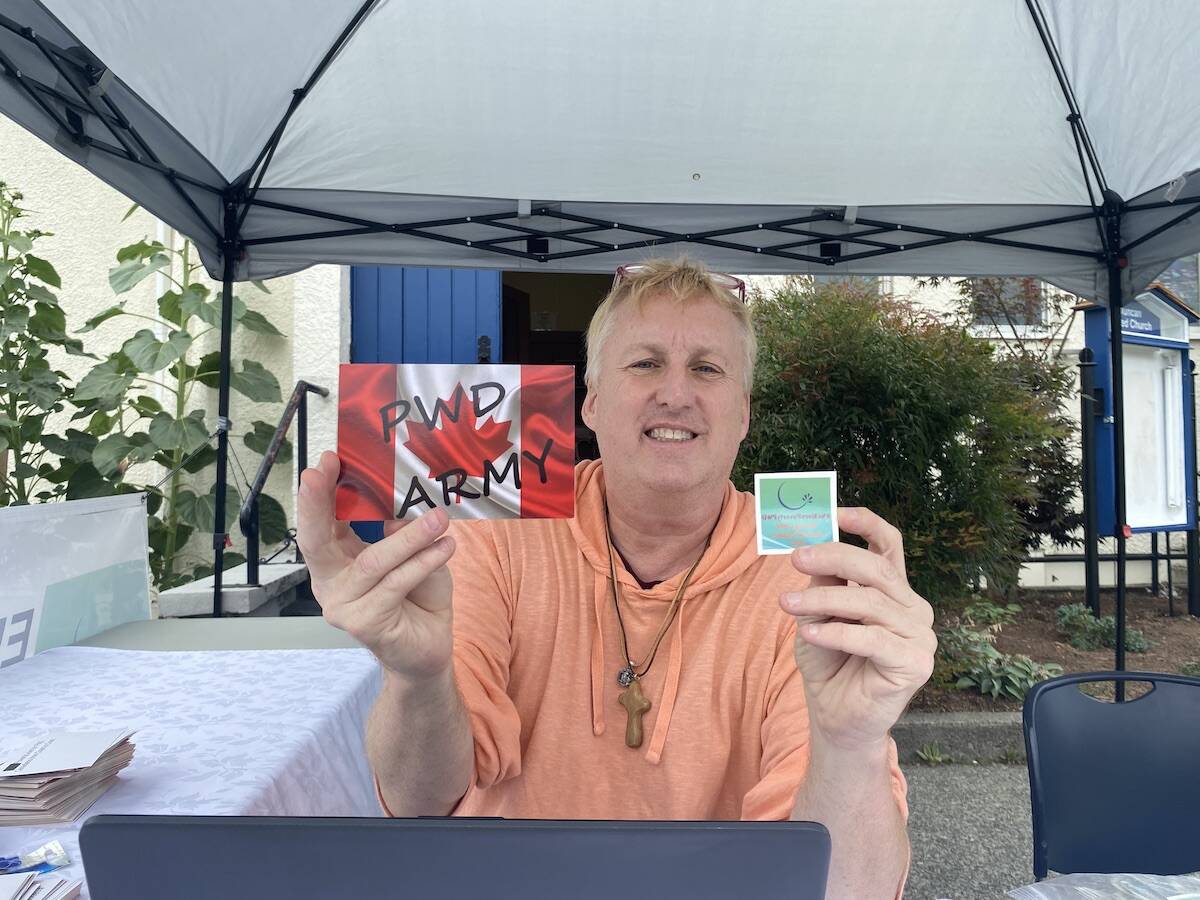 Disability advocate Jeff Leggat of Duncan holds up a postcard which will be mailed to Prime Minister Trudeau, and a temporary tattoo in his efforts to raise awareness about the legislated poverty in Canada and to see it end. Leggat had his booth set up in front of Duncan United Church on Sept. 6 before embarking on his Western Canada tour from Sept. 7 to 16. (Chadd Cawson/Citizen)